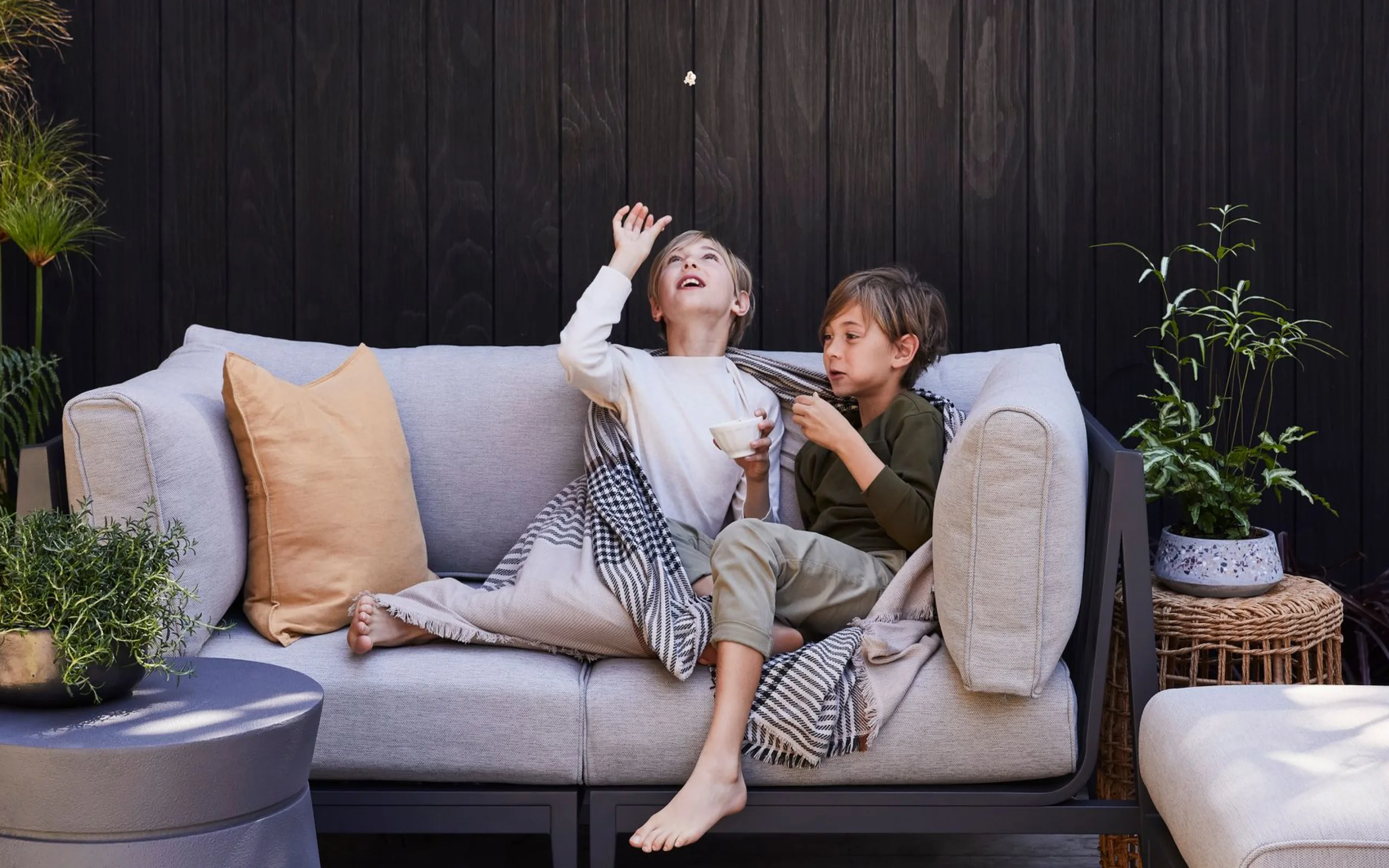 Two children laughing on an outdoor sofa, one tossing popcorn in the air